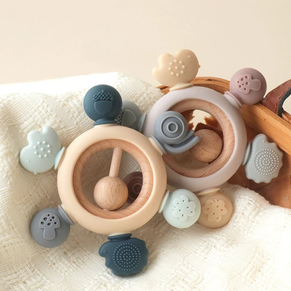 MimiAnneau silicone and wood baby teething rings in pastel blue, beige, and grey tones.